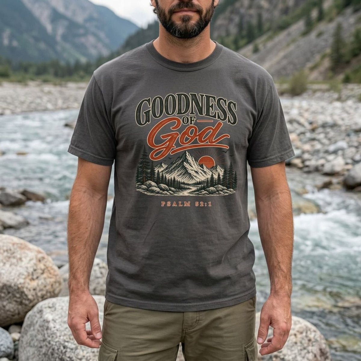 Close-up of a man by a mountain river wearing the Pepper charcoal Goodness of God Psalm 52:1 t-shirt