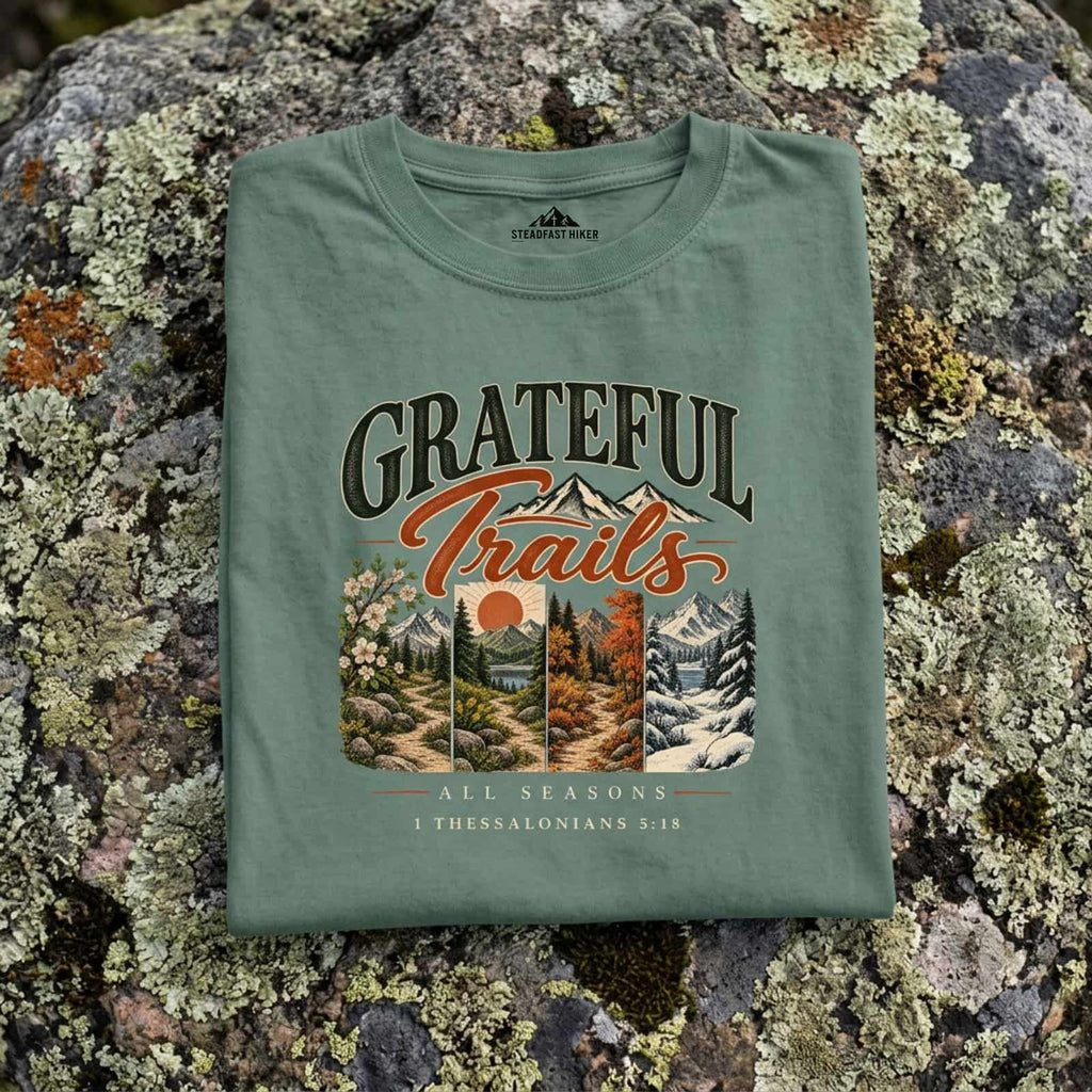 Close-up of the Grateful Trails four seasons design printed on Light green Christian hiking t-shirt resting on a rock, 1 Thessalonians 5:18