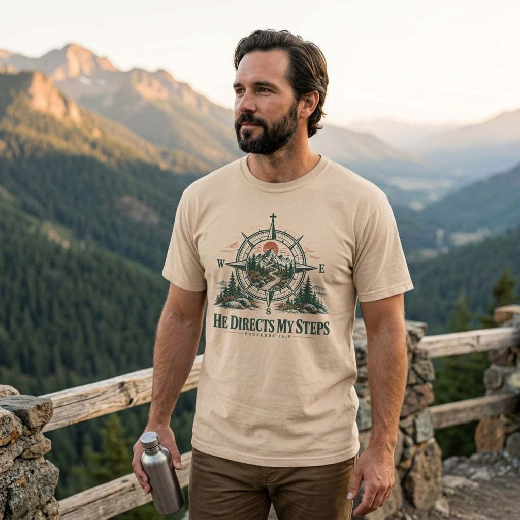 Bearded man at a mountain overlook wearing the Ivory He Directs My Steps t-shirt at sunset