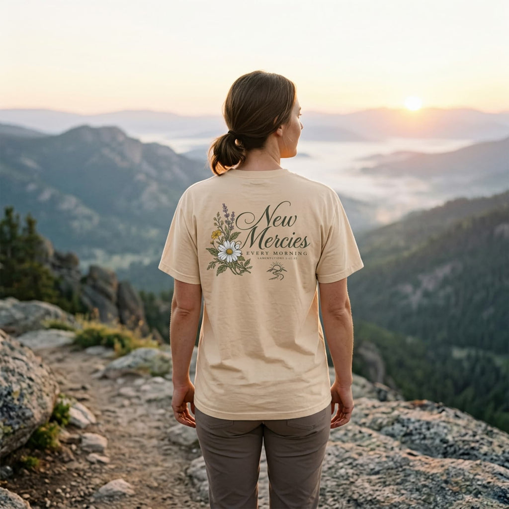 Woman at a mountain overlook at sunrise wearing the Ivory New Mercies Every Morning t-shirt, back print visible