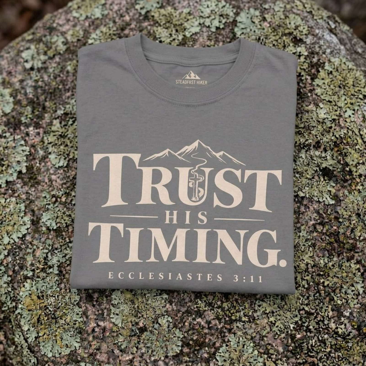 Close-up of the Trust His Timing design printed on a Gray Christian hiking t-shirt resting on a rock, Ecclesiastes 3:11