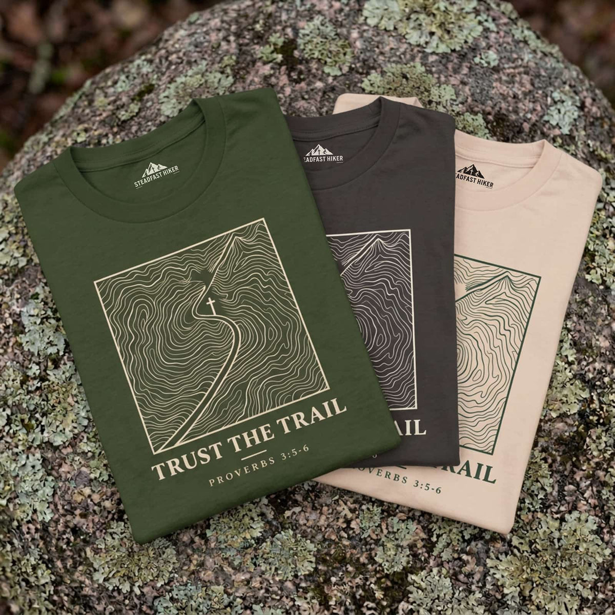 Trust the Trail Christian hiking t-shirts in Sage, Ivory and Pepper laid on a rock